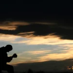 Homem ajoelhado em oração ao entardecer com céu dramático e nuvens douradas ao fundo